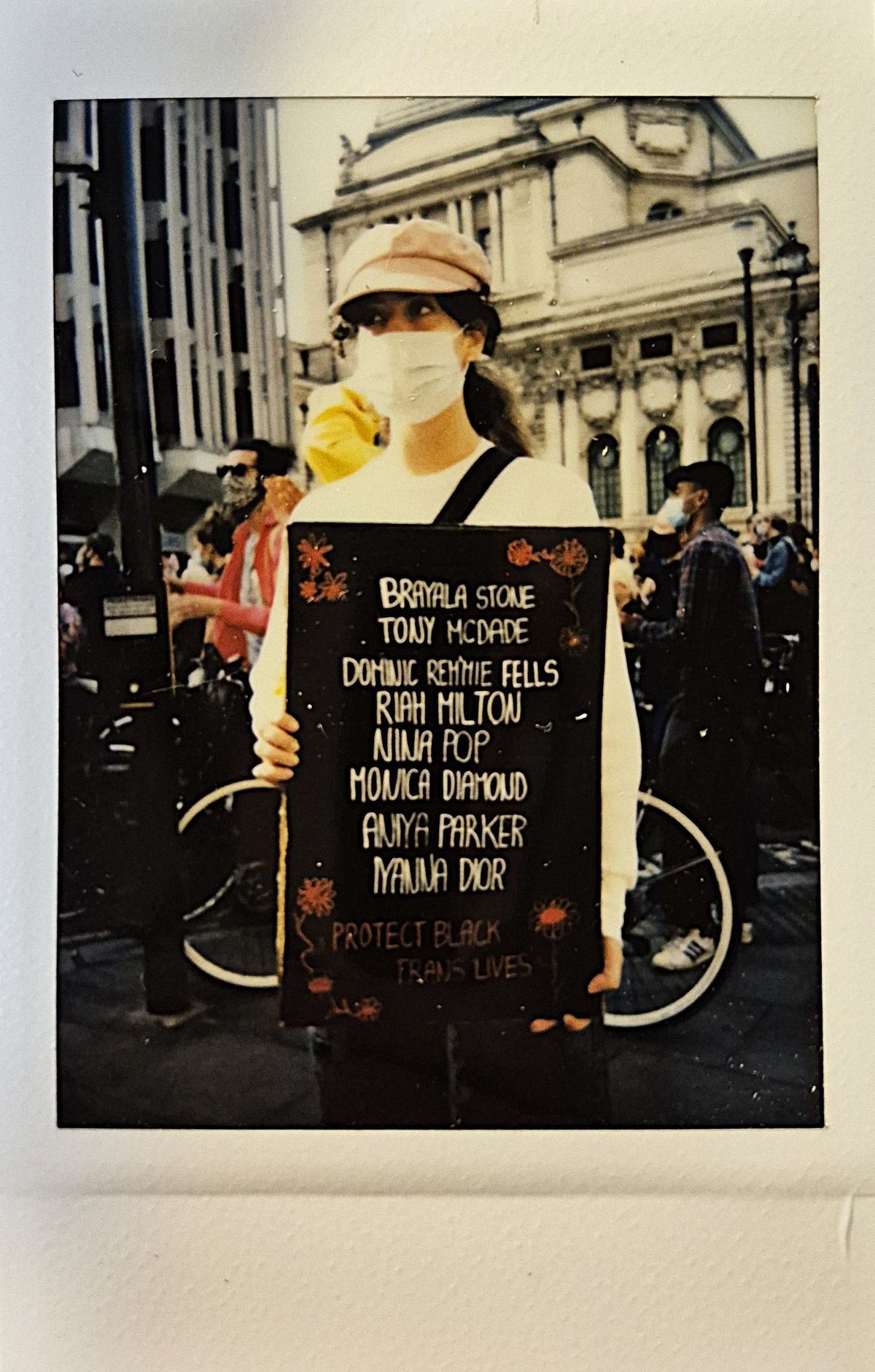 A person wearing a mask holds a sign listing names then "PROTECT BLACK TRANS LIVES".