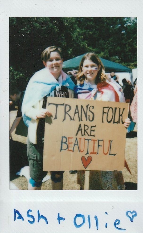 Two people are holding a sign reading "Trans Folk Are Beautiful" with flags draped around them.