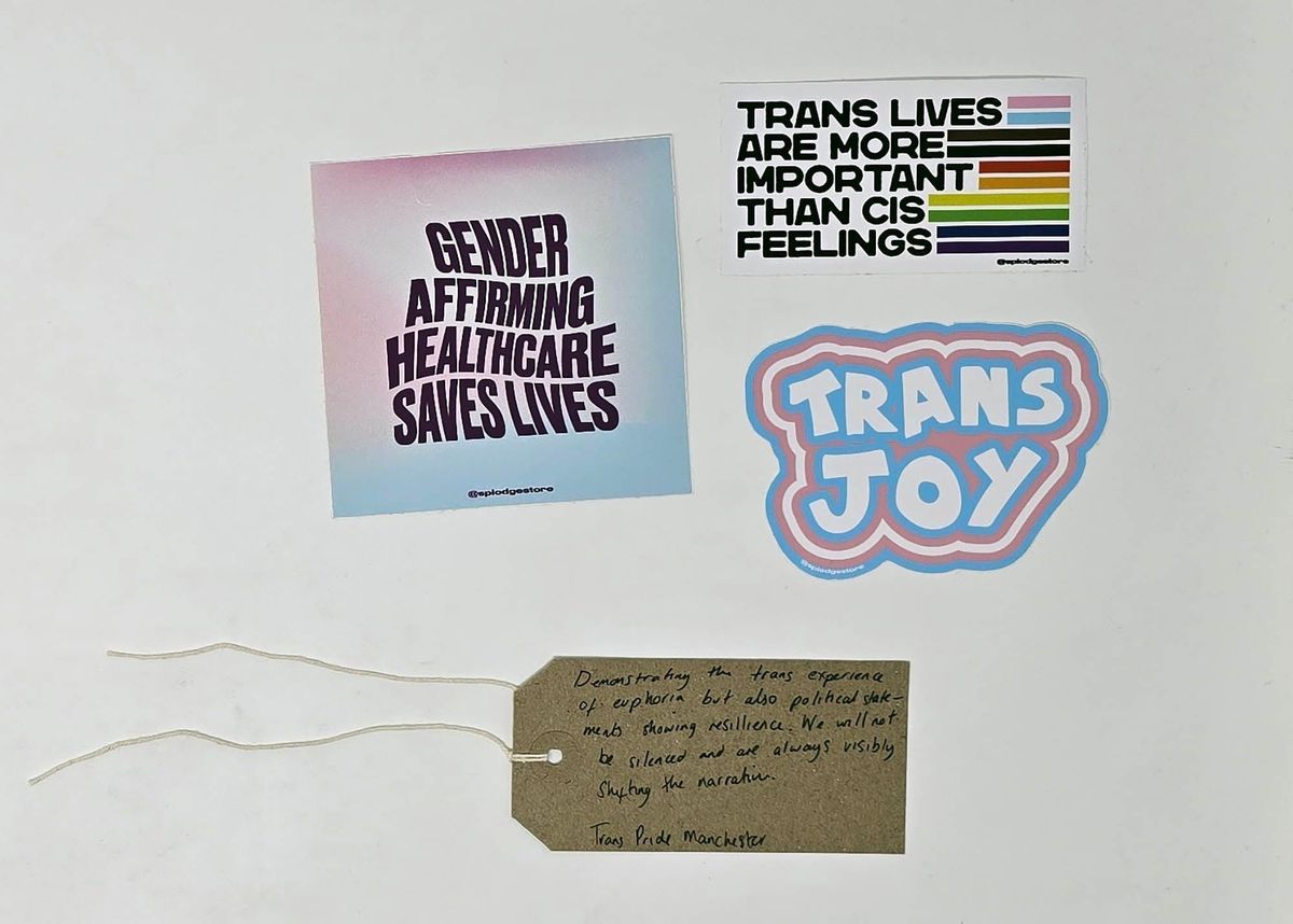 Three stickers and one brown tag laid in a square. Sticker 1 reads "TRANS JOY" and is outlined by trans flag stripes. Sticker 2 reads "TRANS LIVES ARE MORE IMPORTANT THAN CIS FEELINGS" with colourful stripes to the side. Sticker 3 reads "GENDER AFFIRMING HEALTHCARE SAVES LIVES" on a gradient background. The tag reads "Demonstrating the trans experience of euphoria but also political statements showing resilience. We will not be silenced and are always visibly shifting the narrative. Trans Pride Manchester"
