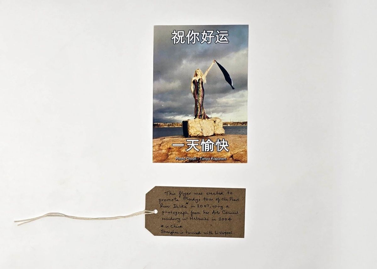 Promotional postcard for Mandy Romero's tour of China. There is Chinese text at the top and bottom and the image is of a person in a dress standing on a rock holding a scarf up in their left hand. With a brown, handwritten luggage tag.