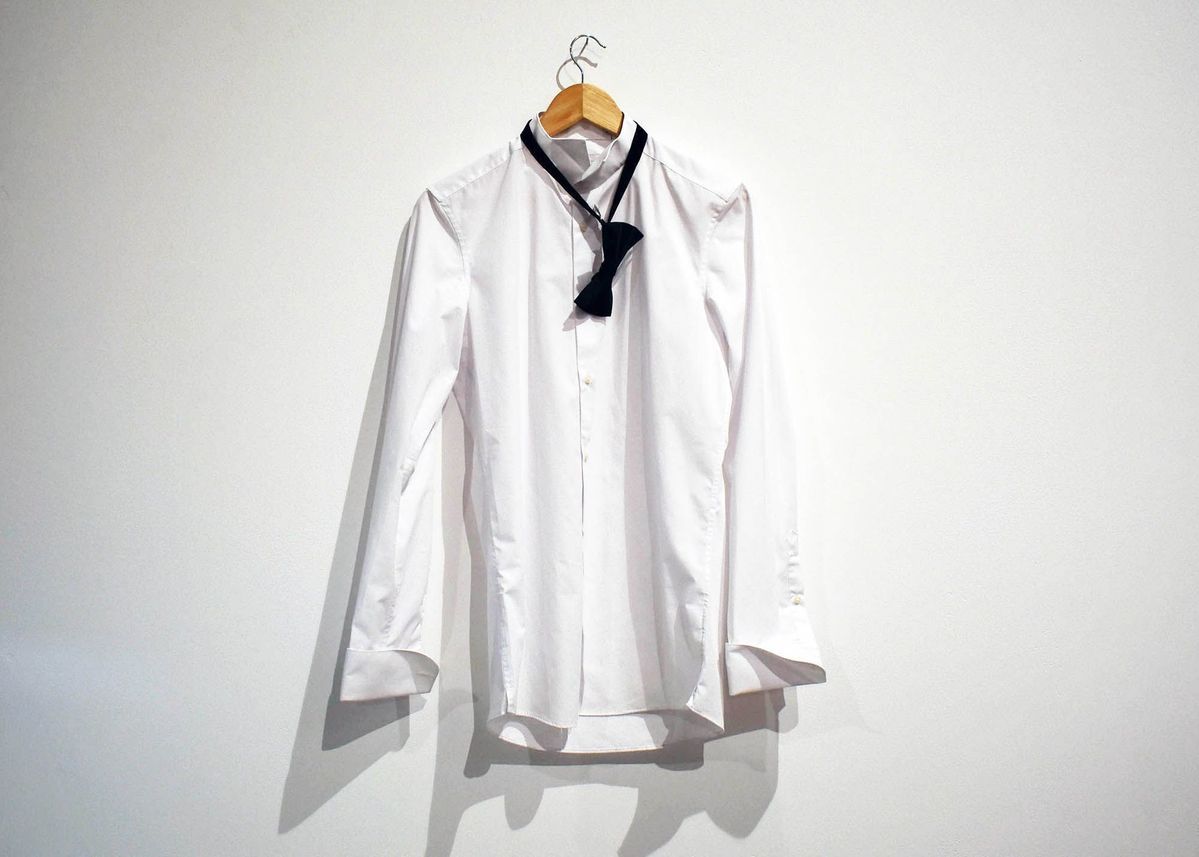 A formal, plain white, long sleeved shirt on a wooden hangar. A black bow tie hangs round the neck.