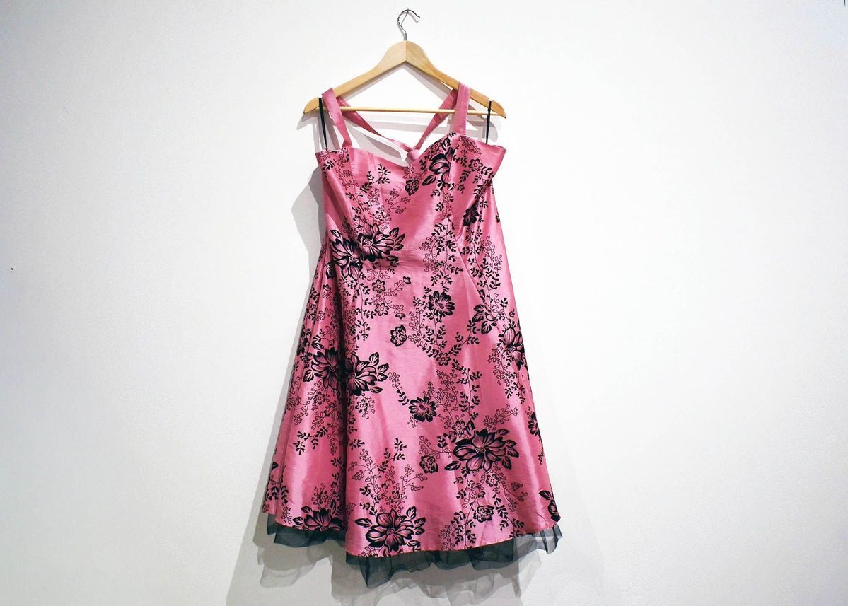 A pink dress with black floral patterns hangs on a wooden hanger against a white wall.