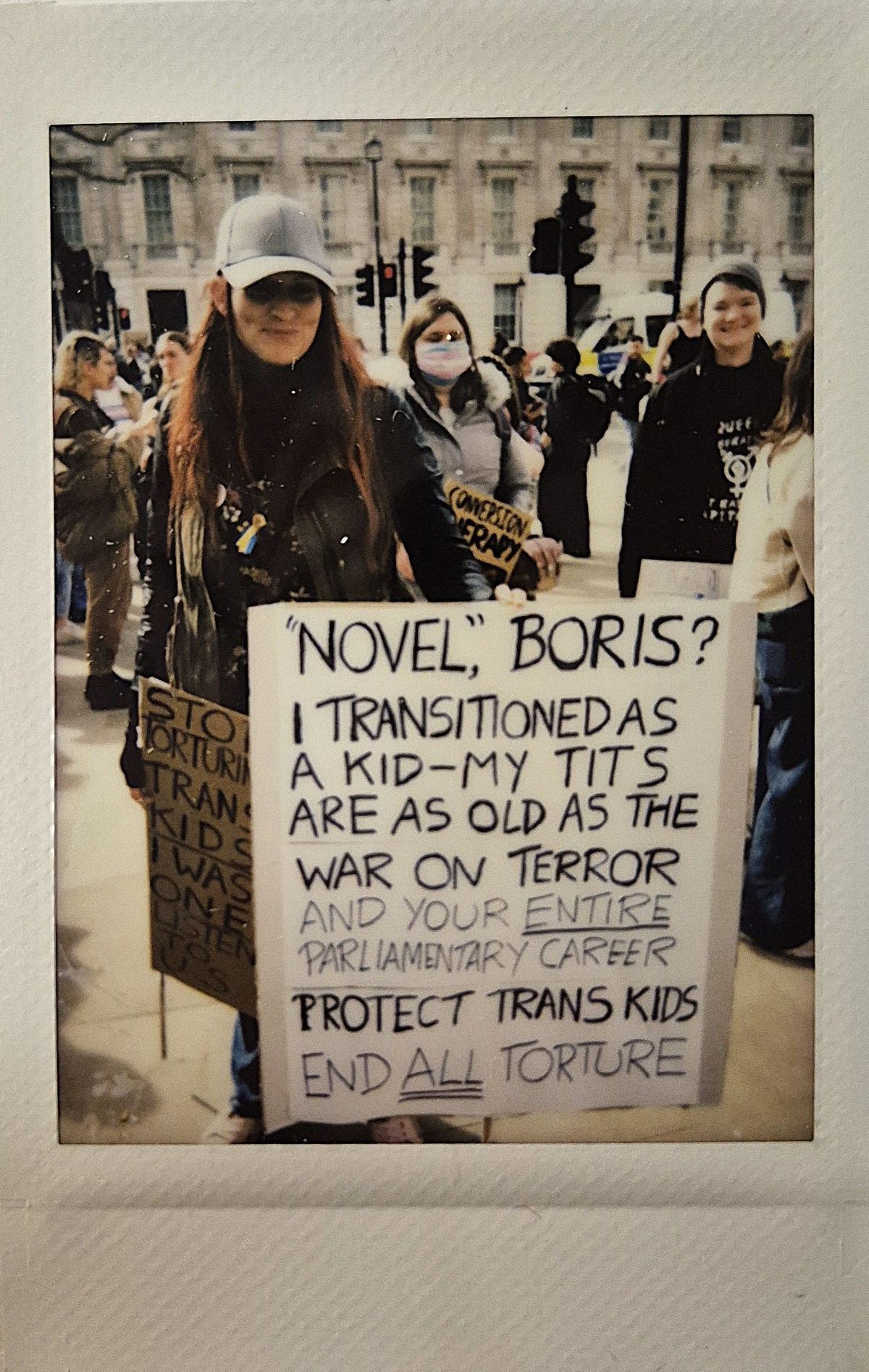 A protester holds a sign which says "NOVEL, BORIS? I TRANSITIONED AS A KID-MY TITS ARE AS OLD AS THE WAR ON TERROR AND YOUR ENTIRE PARLIAMENTARY CAREER PROTECT TRANS KIDS END ALL TORTURE".