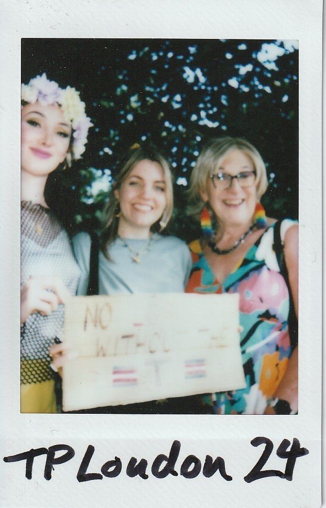Three smiling individuals pose together, one holding a sign, with trees blurred in the background.