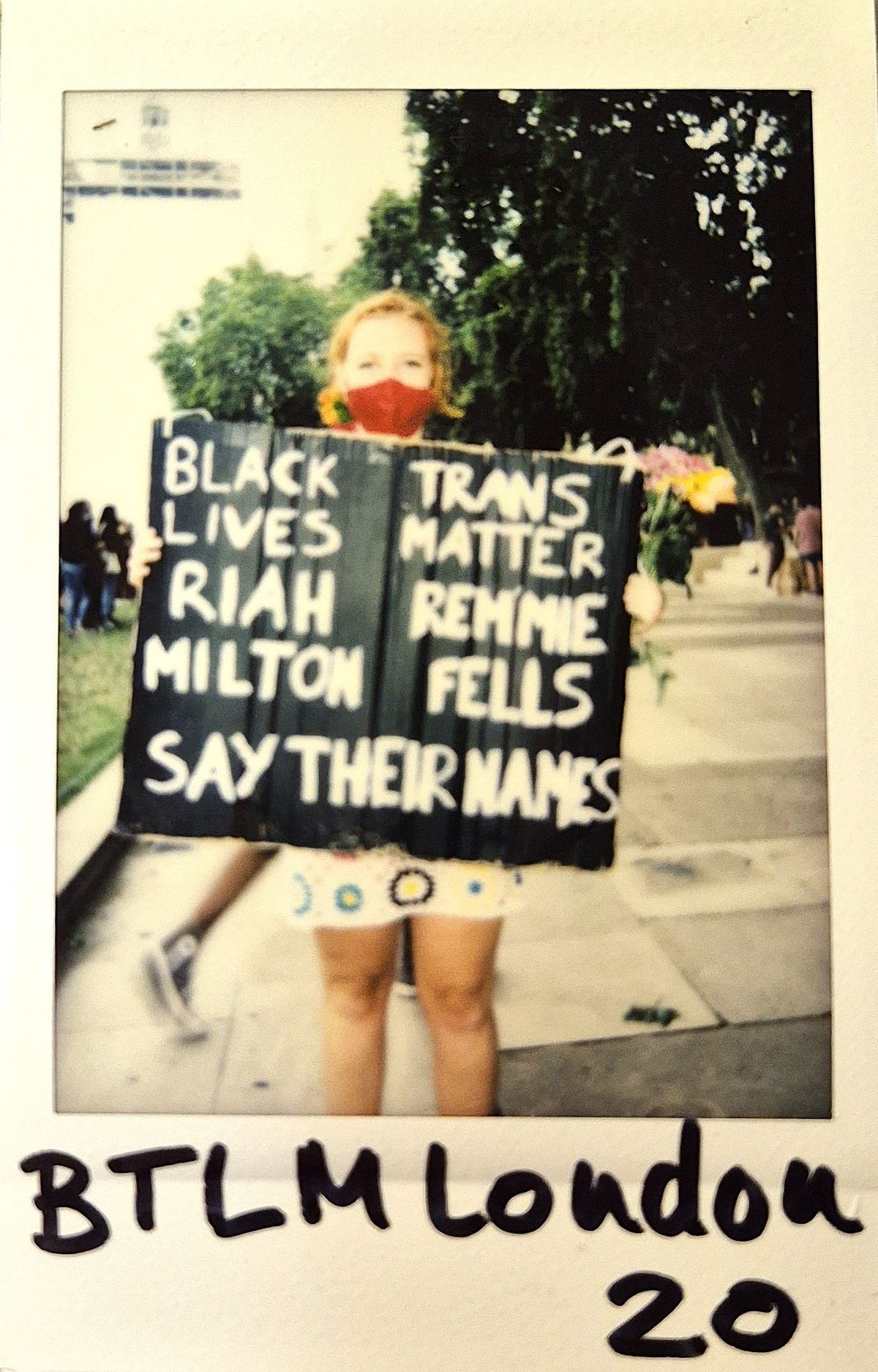 A person wearing a red mask holds a sign advocating Black and Trans Lives Matter.