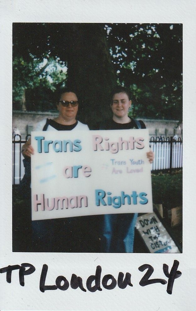 Two individuals hold a sign reading "Trans Rights are Human Rights" during a gathering outdoors.