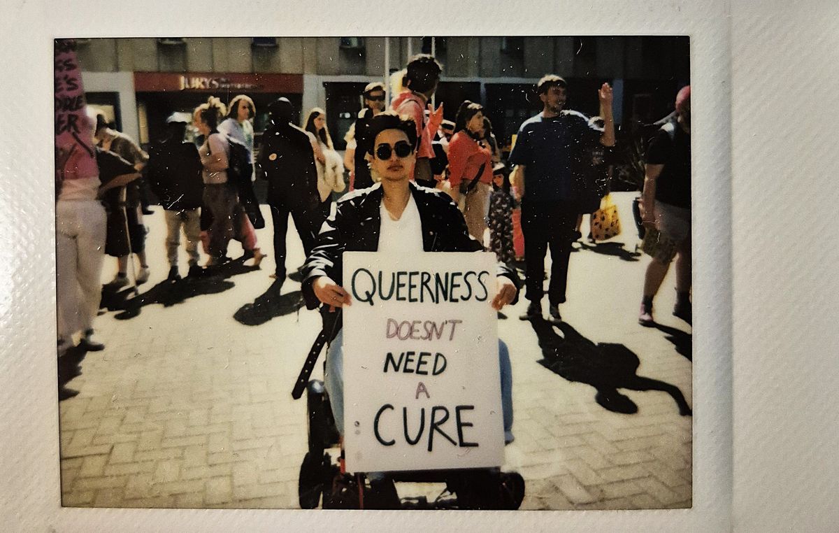 A person holds a sign saying "Queerness doesn't need a cure" whilst they are sat in their wheelchair.
