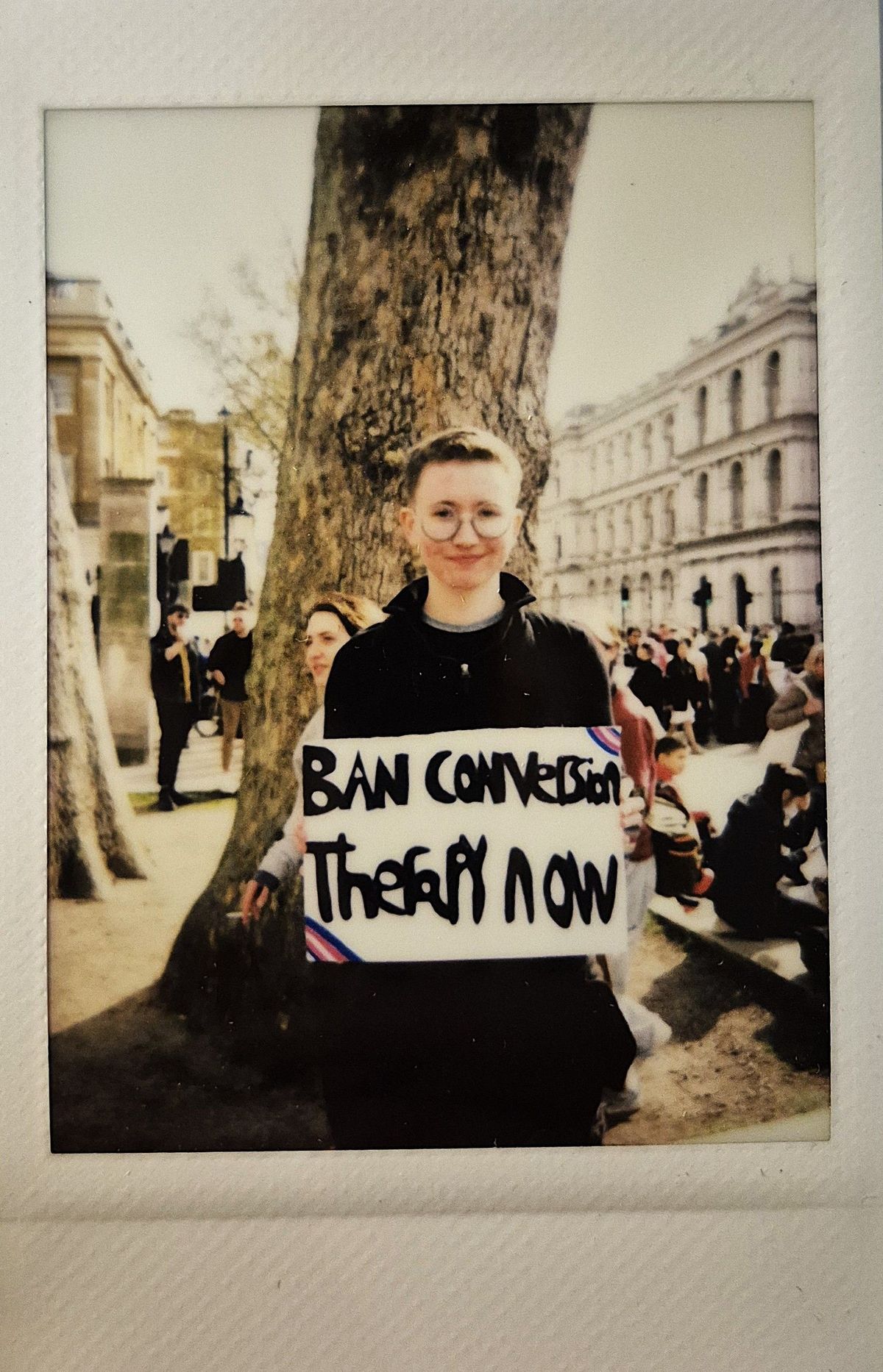 A person holding a sign reading "Ban Conversion Therapy Now" stands near a large tree.