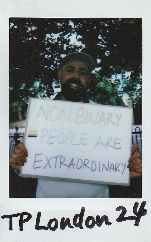 A person holds a sign saying "Non-Binary People Are Extraordinary" with trees in the background.