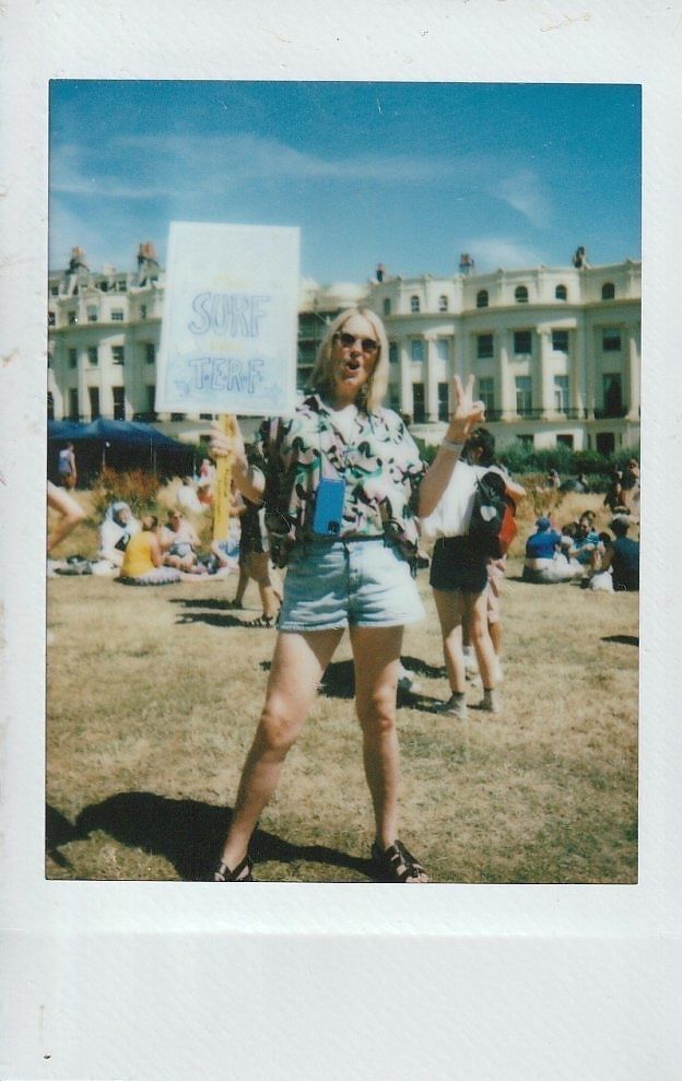 A person holding a "Surf the Turf" sign poses joyfully in a sunny, crowded outdoor gathering.