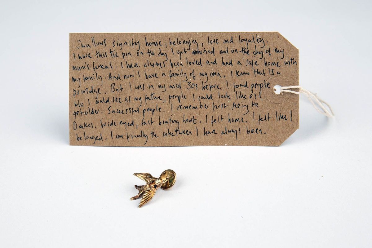 A small golden pin which is a swallow. Behind it is a brown luggae tag with writing on it.