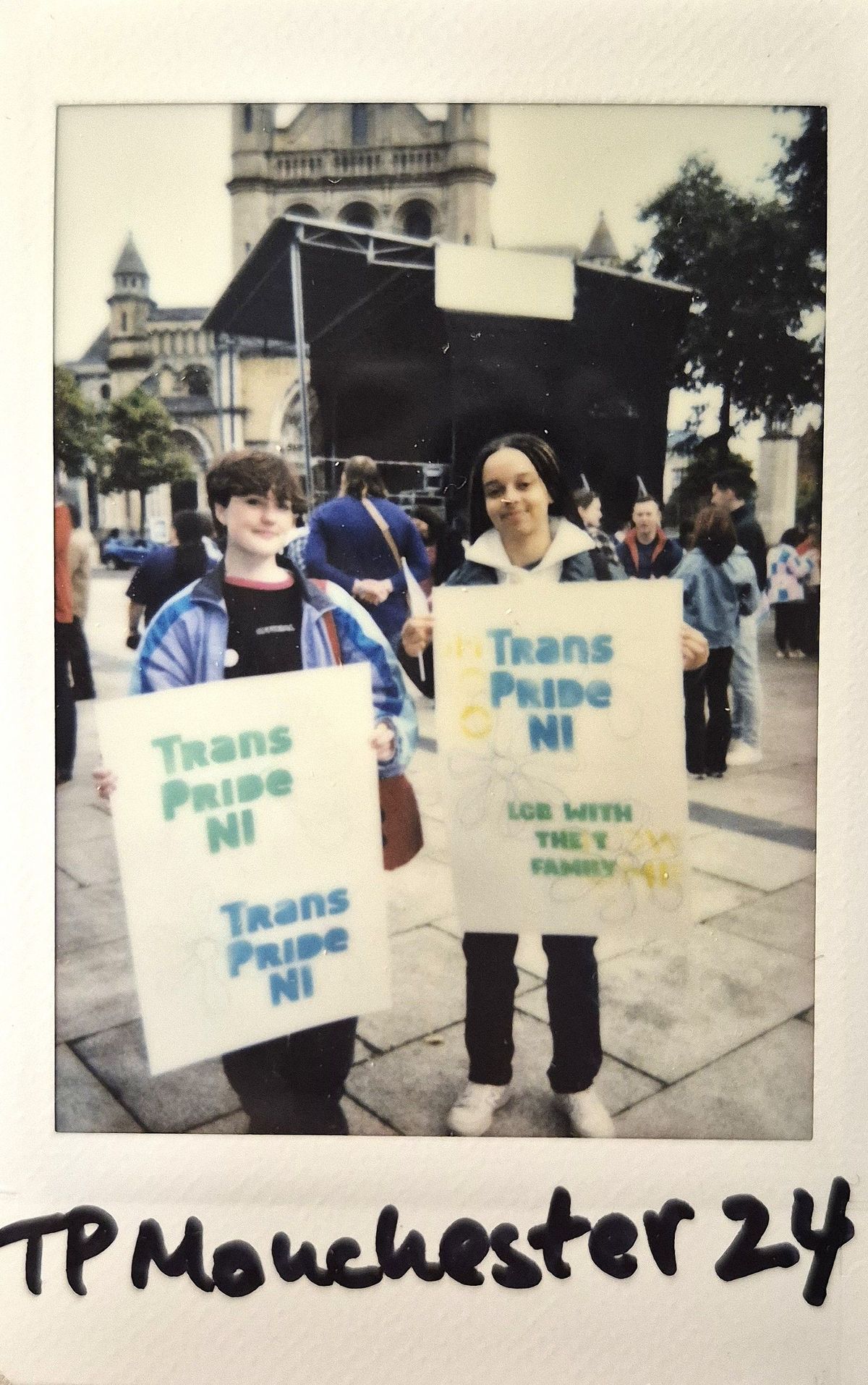 Two people stand holding Trans Pride NI signs, with a historic building in the background.