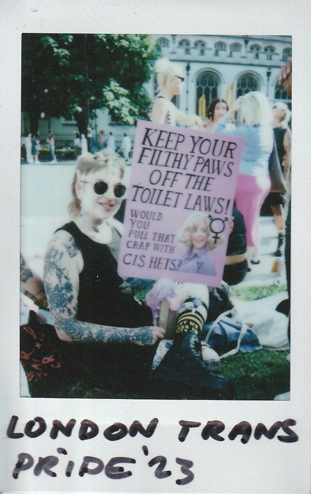 A person sat on the grass holding a placard which says "Keep Your Filthy Paws Off The Toilet Laws".