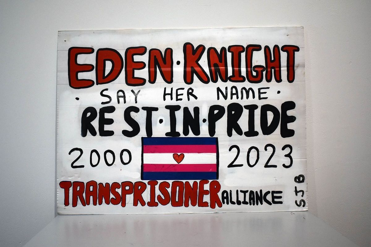 A white plastic placard reading "EDEN KNIGHT SAY HER NAME REST IN PRIDE 2000 - 2023 TRANSPRISONER ALLIANCE SJB" in red and black, handwritten text. Between the dates, there is a trans flag with a heart in the centre.