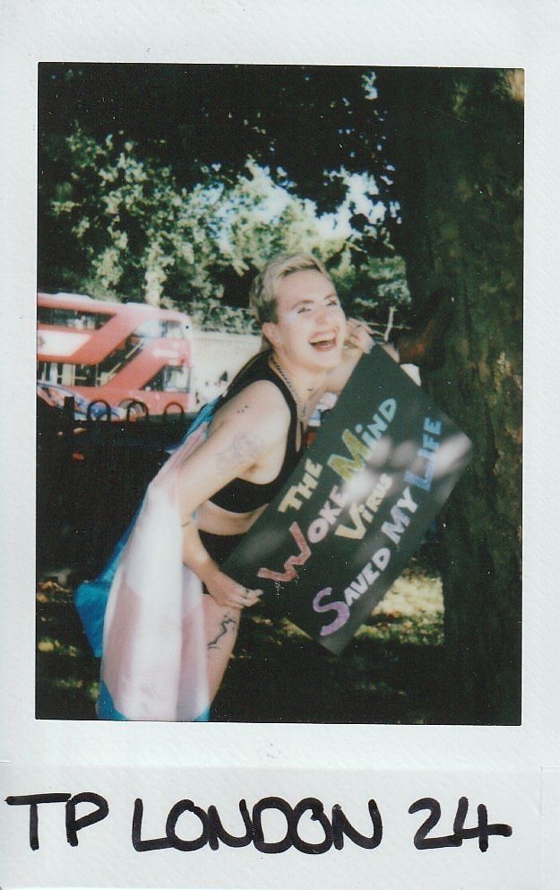 A smiling person stands under a tree, holding a colorful sign with text, next to a red bus.