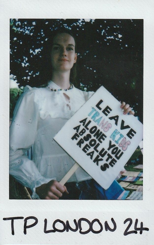 A person holding a sign advocating for trans kids under a tree in London setting.