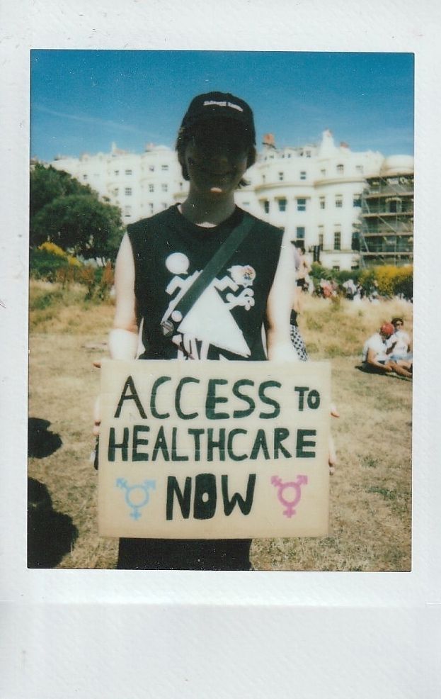 A person holds a sign reading "ACCESS TO HEALTHCARE NOW" during a sunny outdoor protest.