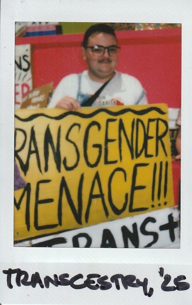 A person with glasses smiles while holding a yellow sign with bold text reading, "TRANSGENDER MENACE".