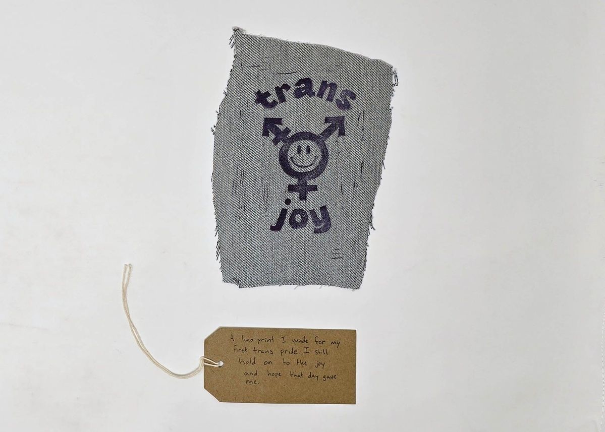 A lino cut print on a scrap of blue denim. Text "trans joy" around the trans logo (stylised with a smiley face in the centre). With a brown handwritten luggage tag.