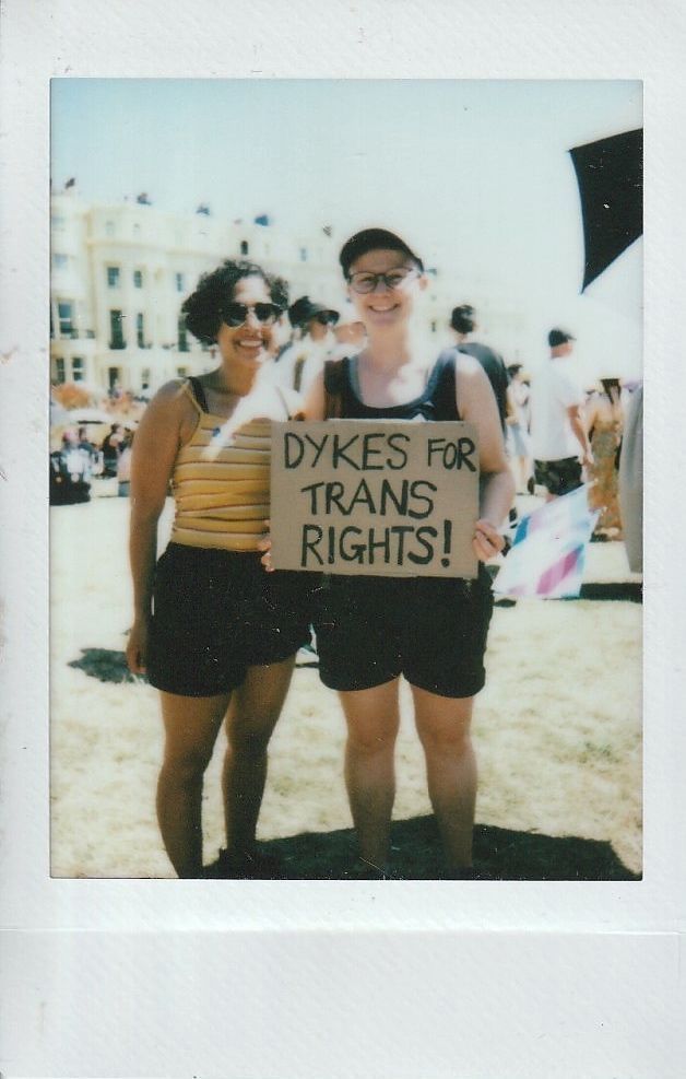 Two individuals stand together holding a sign which says "DYKES FOR TRANS RIGHTS".