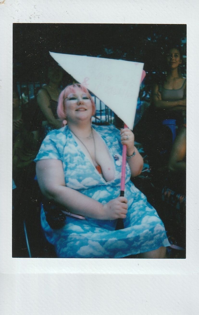 A person with pink hair smiles while holding a large triangular sign, surrounded by others.