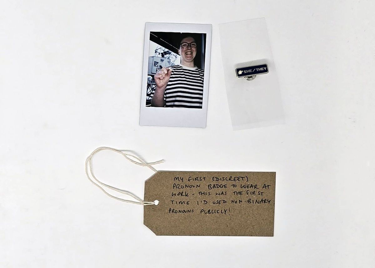 A Polaroid of a person in a striped top wearing glasses and holding a small badge, a blue enamel 'SHE/THEY' pin badge and a handwritten brown luggage tag.