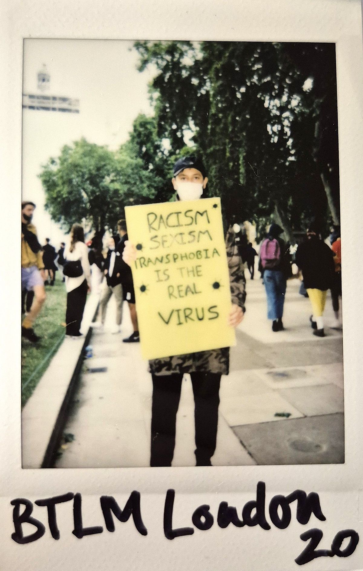 A person holds a yellow sign reading "Racism, Sexism, Transphobia Is the Real Virus".