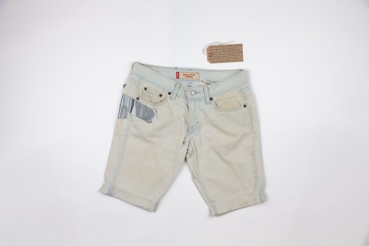 Front of a pair of white shorts with the cuffs rolled up. There is a small tear next to the fly of the shorts and duct tape visible on the right hand side which is wrapping around from the back of the shorts.