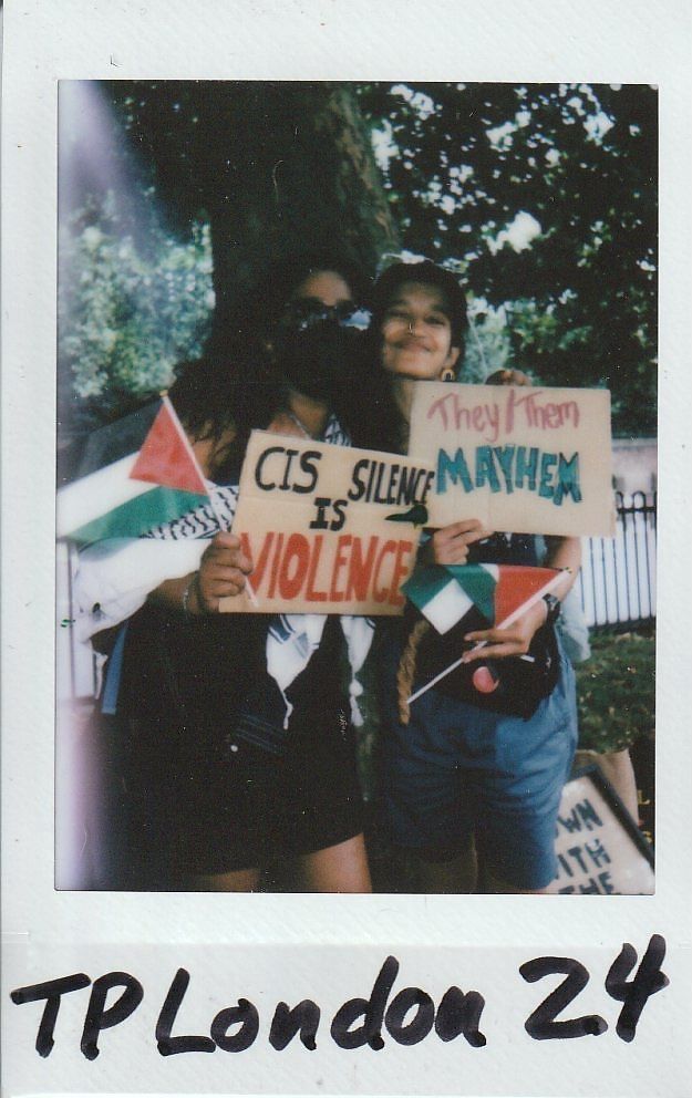 Two people hold protest signs, one reads "Cis Silence is Violence," and the other says "They/Them Mayhem" under a tree.