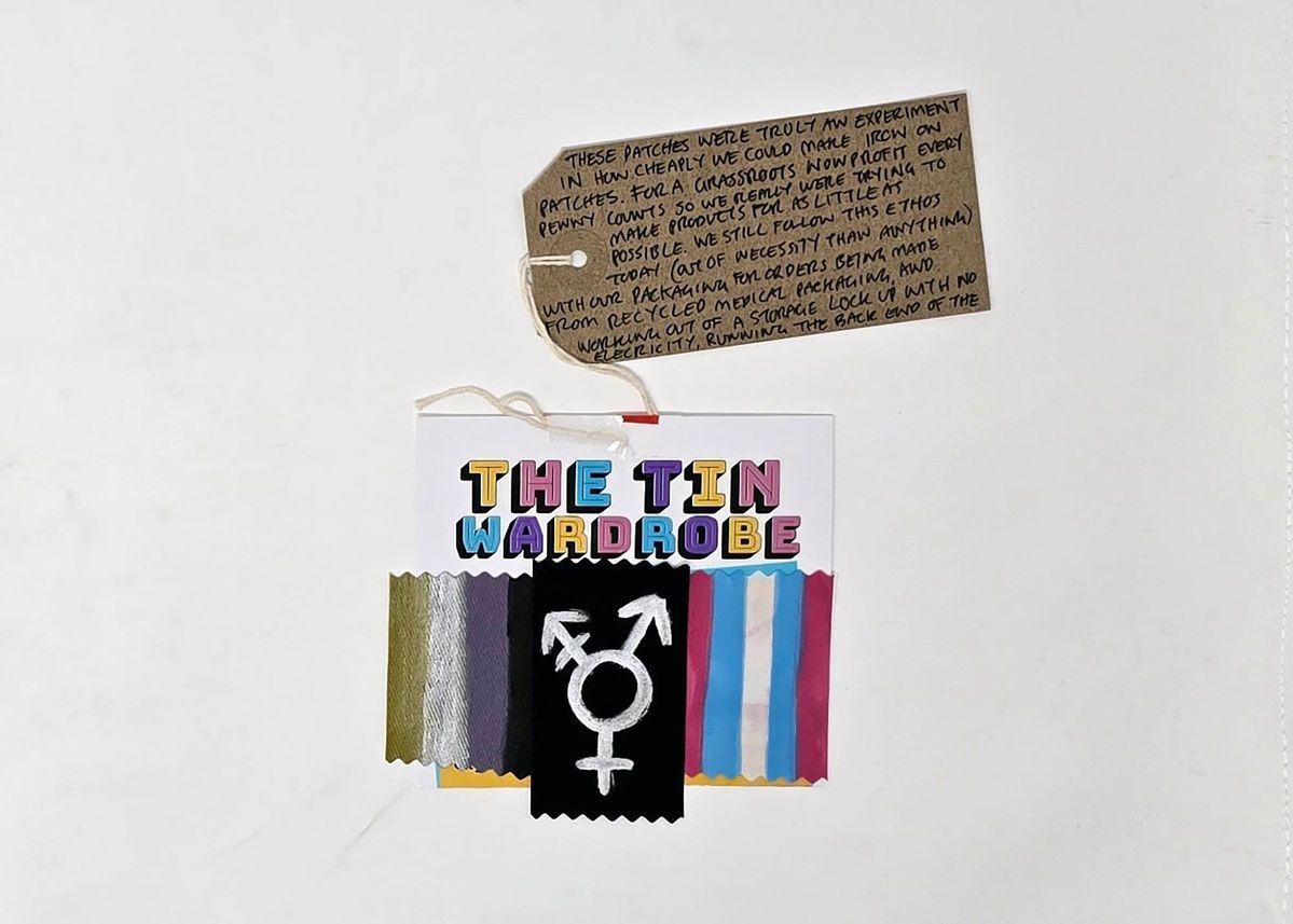 The fabric patches depicting the Non-Binary Flag, Trans Flag and the Trans Symbol, mounted on a card titled "THE TIN WARDROBE", with a brown handwritten luggage tag.