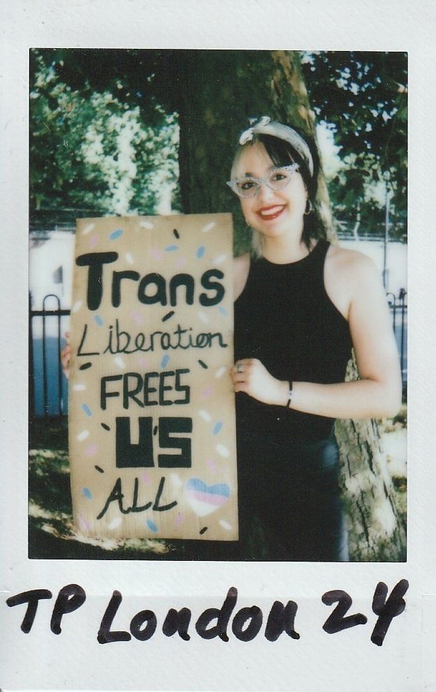 A person stands by a tree, smiling, holding a sign saying, "Trans Liberation Frees Us All".