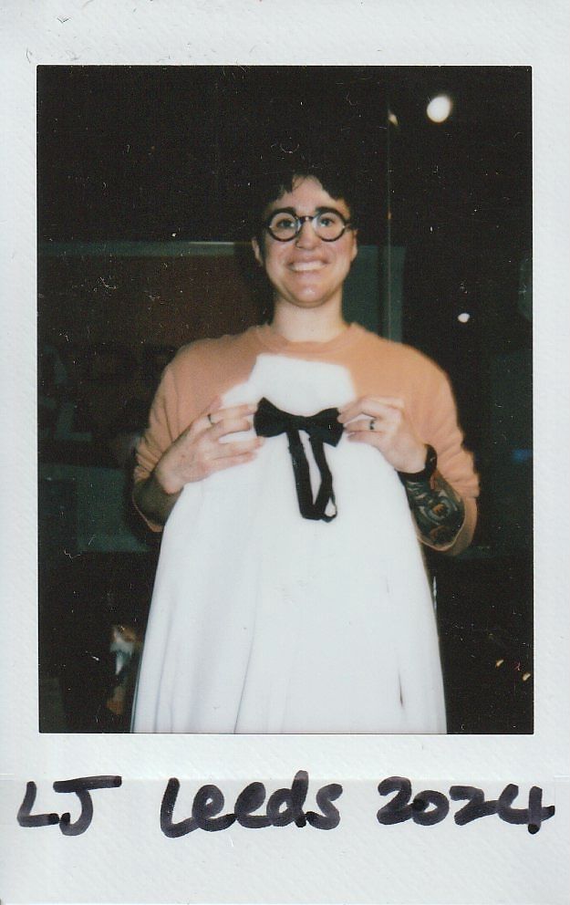 A smiling person stands holding a white garment with a black bow in their hands inside a room.