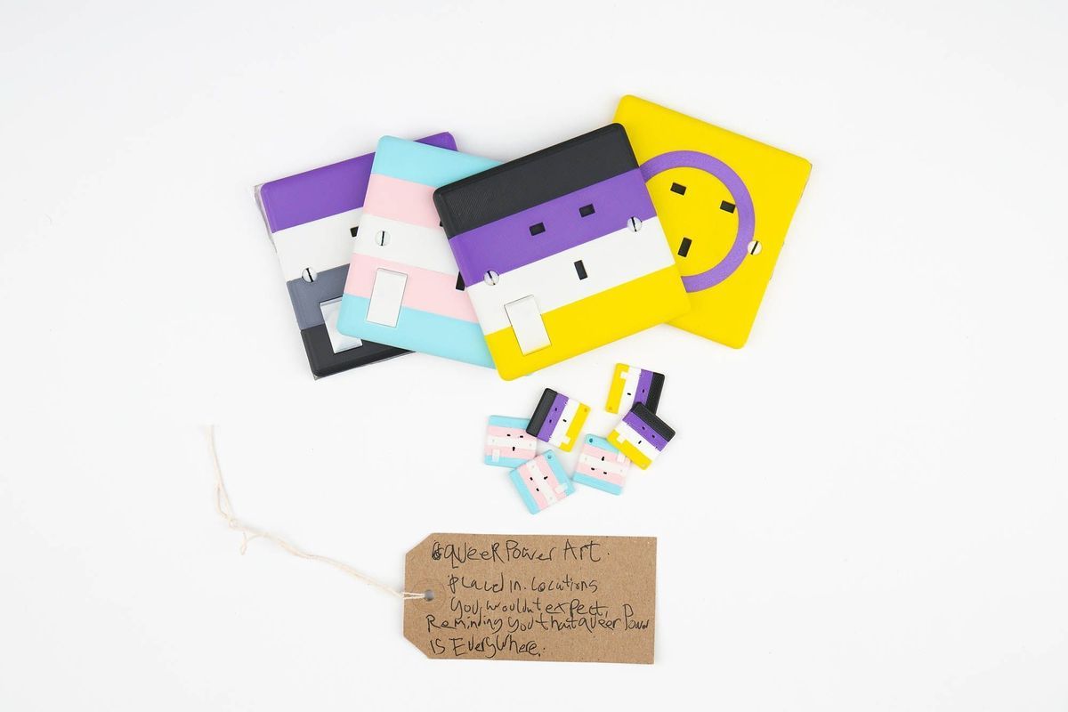 Several 3D printed 3-pin socket facias coloured with the Asexual, Trans, Non-Binary and Intersex flag colours. With a brown, handwritten luggage tag.