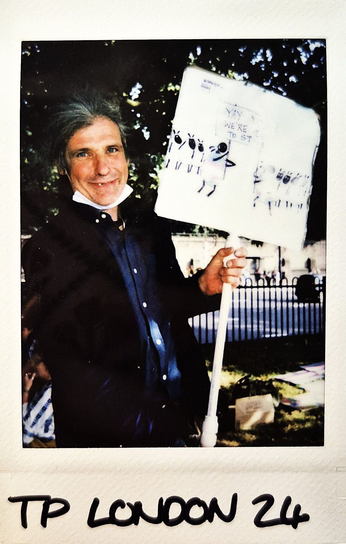 A man stands outdoors holding a protest sign with sheep illustrations, smiling confidently at the camera.