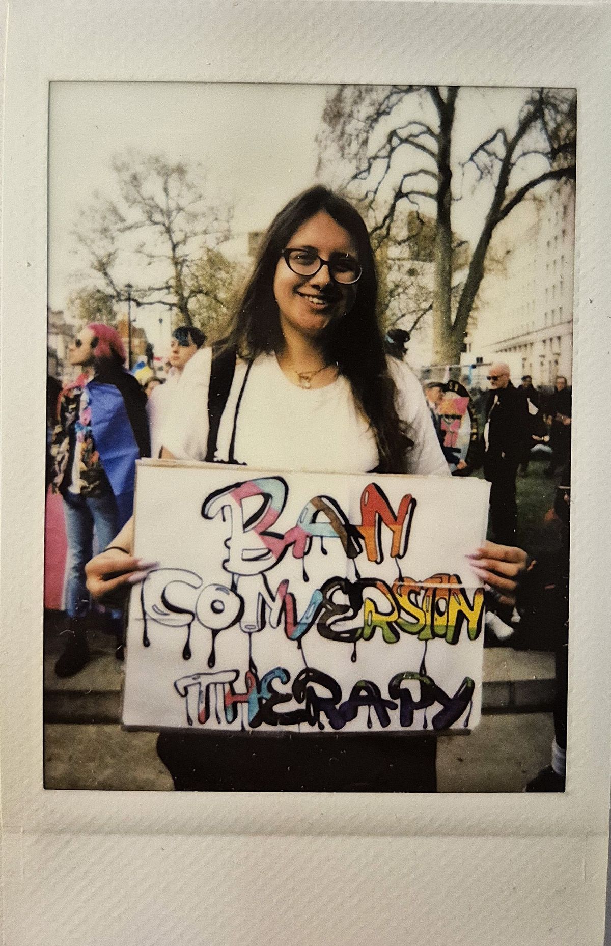 A person holds a colorful sign reading "Ban Conversion Therapy" in a busy outdoor setting.