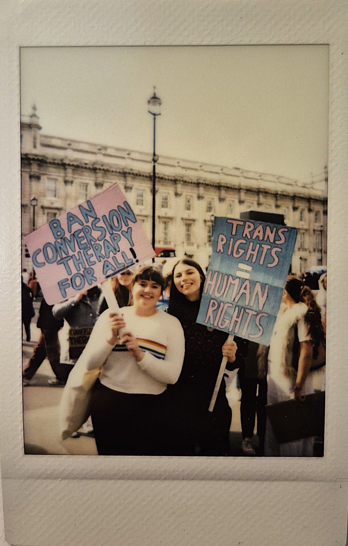 People are holding colorful protest signs one which says "BAN CONVERSION THERAPY FOR ALL!" and the other "TRANS RIGHTS = HUMAN RIGHTS".