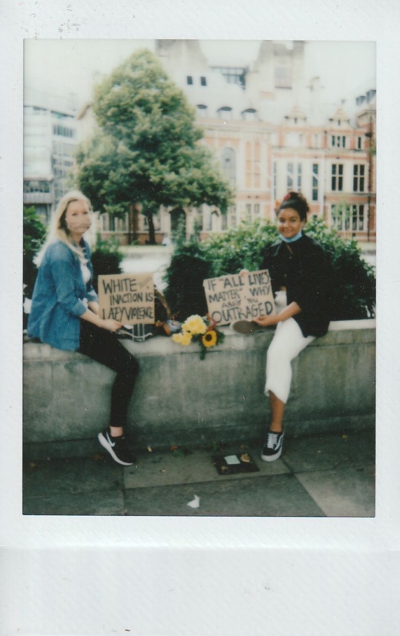 Two people sit on a low wall, holding signs which say "White inaction is lazy violence" and the other says "If 'all' lives matter why are you outraged".