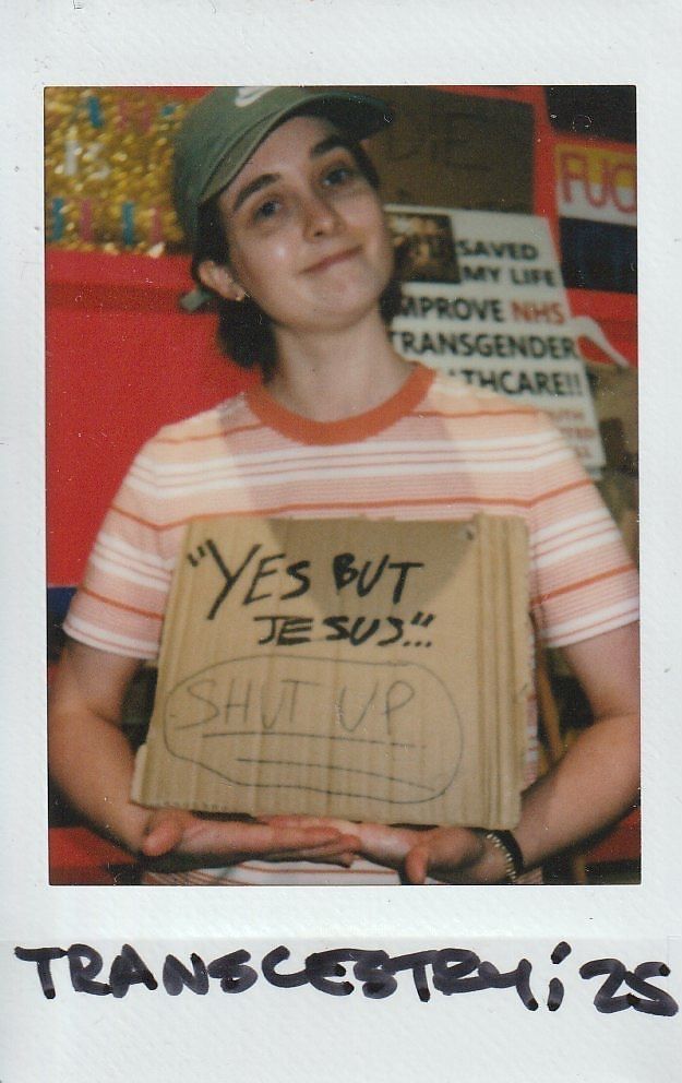 A person wearing a cap holds a cardboard sign with handwritten messages, smiling gently at the camera.