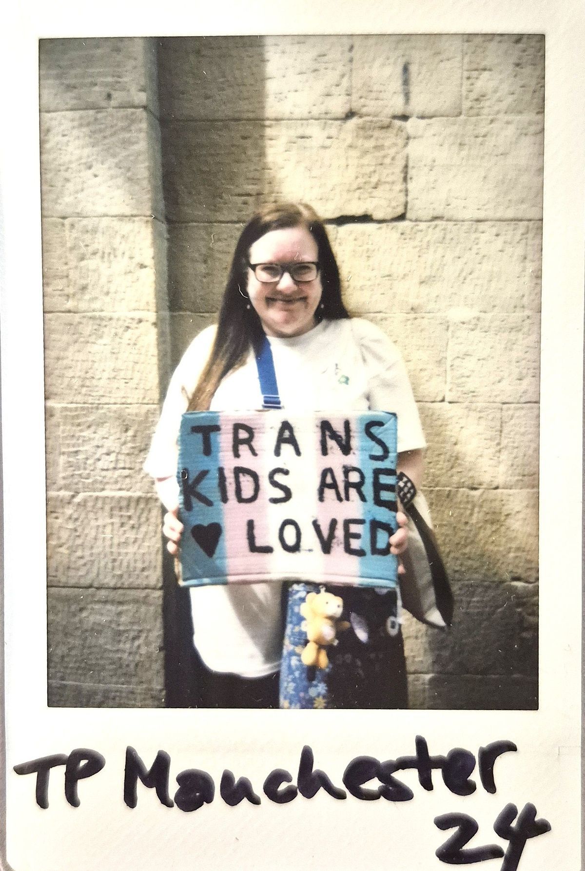 A smiling person holds a colorful sign reading "TRANS KIDS ARE LOVED" against a stone wall