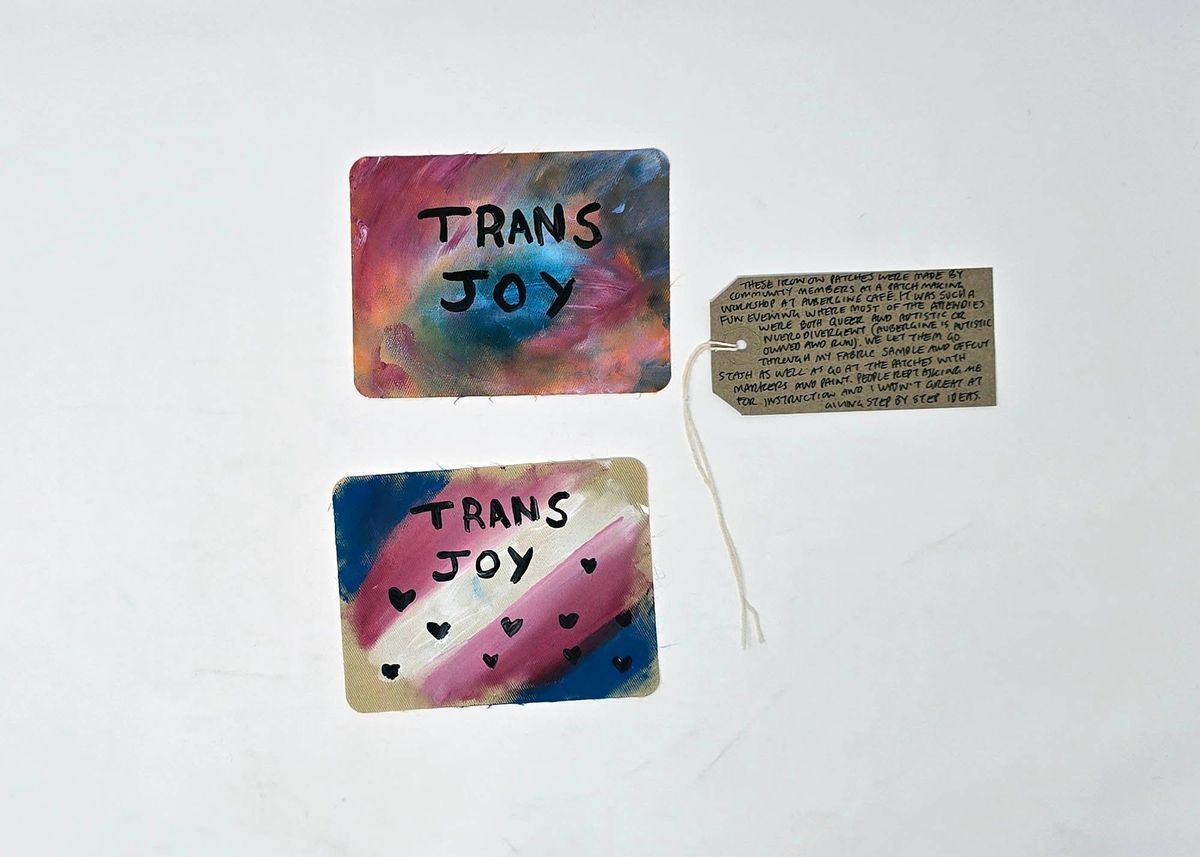 Two colourful fabric patches, one in the trans flag colours, both with "TRANS JOY" written on them.  With a brown, handwritten luggage tag.