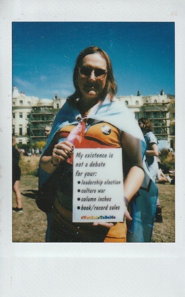 A person wearing sunglasses and a cape holds a sign which says "My Existence Is Not A Debate For Your -leadership election -culture war -column inches -book/record sales #NotSafeToBeMe'.