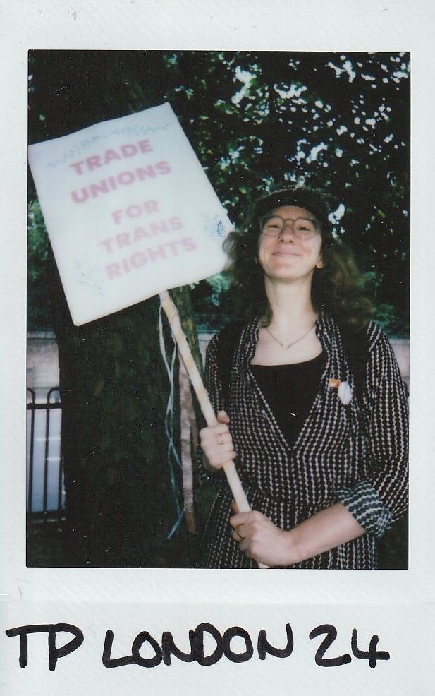 A person is standing outside holding a sign that reads "Trade Unions for Trans Rights" and smiling.