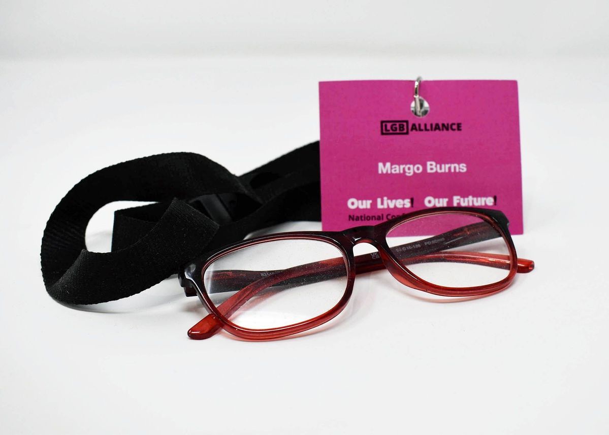 A photograph of a pair of red glasses resting next to a pink name badge which says 'LGB Alliance' with a black lanyard.