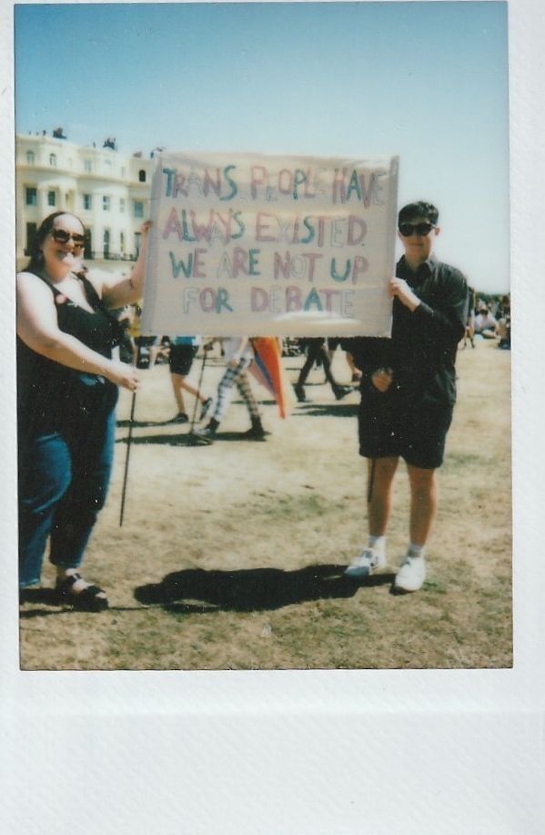 Two individuals hold a sign that says "Trans people have always existed. We are not up for debate".