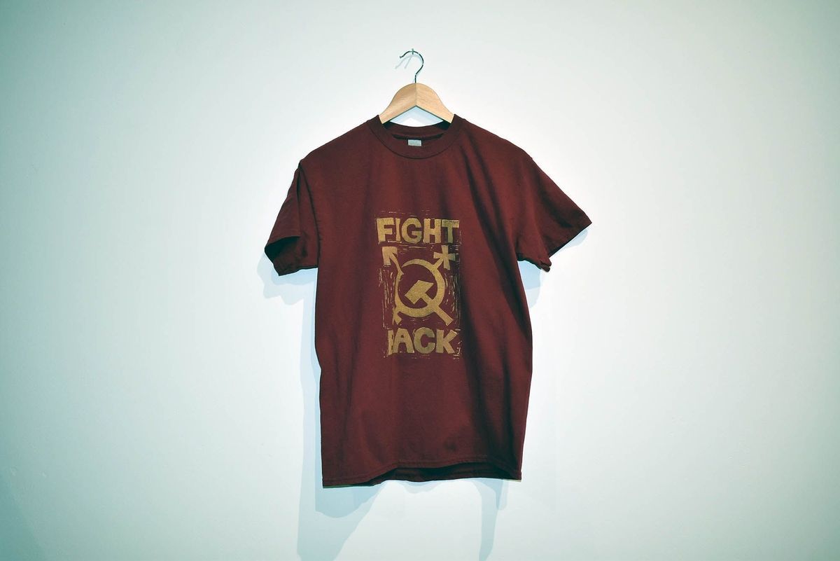 A red t-shirt printed in gold with the trans symbol stylised as a hammer and sickle and the text "FIGHT BACK". On a wooden hanger.