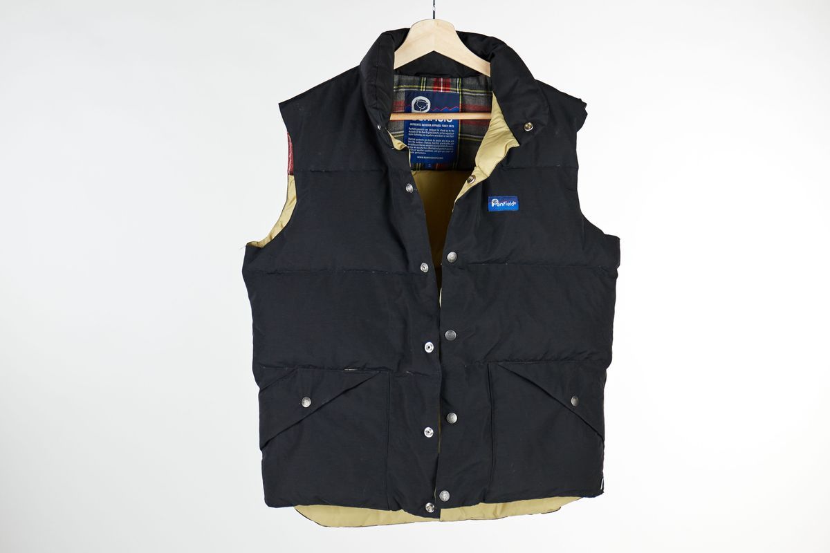 A black Penfield gilet with a cream and tartan lining and poppers down the front and on the pockets on a wooden hangar.