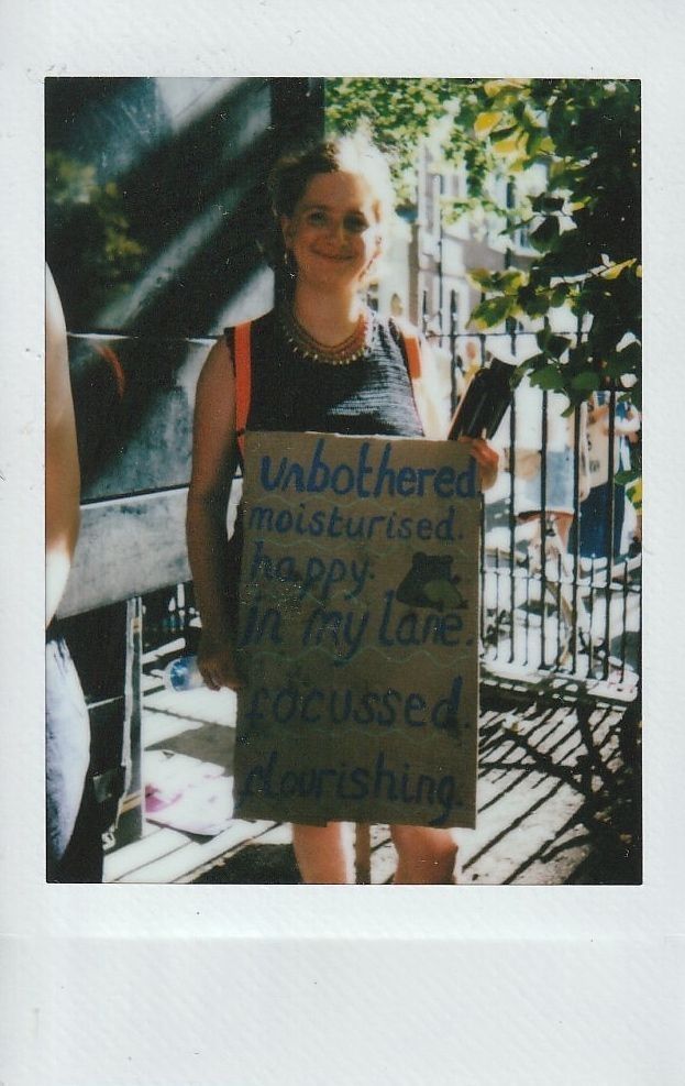 A person stands outside holding a placard which says "Unbothered, moisturised, happy, in my lane, focused, flourishing".