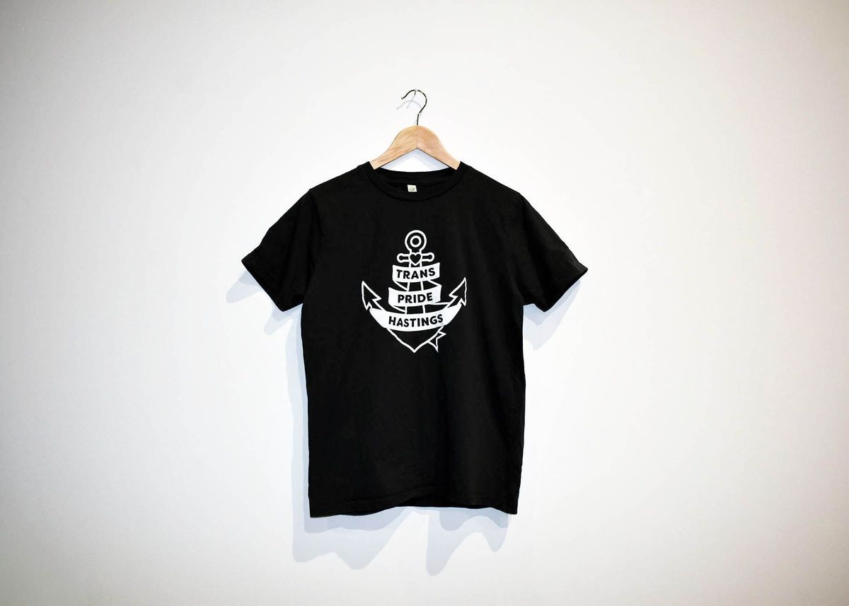 A black T-shirt with "Trans Pride Hastings" is hanging on a wooden hanger.