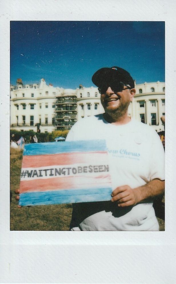 A person holds a colorful sign with "#WAITINGTOBESEEN" outdoors by a large building.