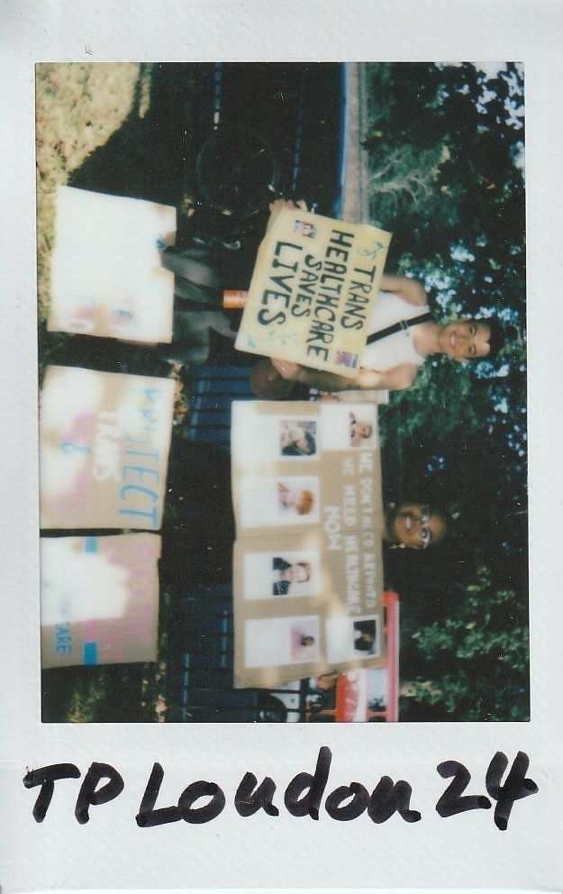 Two individuals hold signs advocating for trans healthcare, standing outdoors with trees behind them.